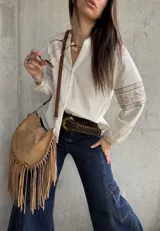 Camisa de inspiración boho de color crudo, con mangas largas abullonadas y puños decorados con detalles bordados geométricos en tonos tostados y dorados. Presenta cuello en V con cierre frontal de botones.