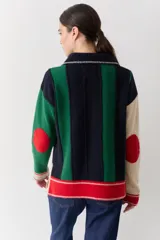 Buzo de punto con cuello polo y cierre frontal de tres botones. Presenta un diseño a rayas verticales en tonos beige y marrón, con mangas en bloques de color rojo y detalles en rosa en los puños y el ruedo.