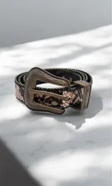 Cinto con estampado de piel de serpiente en tonos marrones, con hebilla y pasador de metal en color bronce envejecido con detalles grabados. Presenta bordes decorados con pequeñas tachas metálicas.