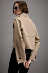 Trench corto de color beige con cuello de solapa, cierre frontal cruzado con botones y mangas largas con trabillas ajustables en los puños. Presenta un diseño estructurado con hombreras y bolsillos delanteros de vivo.