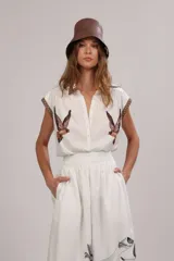 Camisa blanca de poplin de algodón con cuello camisero, abotonadura frontal y mangas cortas. Presenta un estampado de aves rapaces en el frente y galones decorativos en tonos marrones y beige en cuello y mangas.