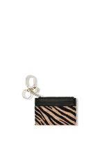 Tarjetero con cierre de cremallera y cuatro ranuras externas para tarjetas. Presenta un diseño con estampado animal print tipo cebra, detalles en material sintético negro y herrajes metálicos dorados, incluyendo una anilla y un clip para llavero.