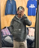 Rompeviento gris de estilo canguro con capucha, cuello alto y bolsillo frontal tipo sobre con cierre. Presenta puños elásticos y diseño funcional impermeable.