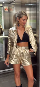 Chaqueta de estilo camisero en color dorado metalizado, con cuello clásico, cierre frontal con botones, mangas largas y bolsillos frontales con solapa.