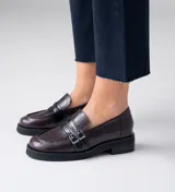 Mocasín de cuero vacuno negro con detalle de pieza de cuero con textura de cocodrilo y hebillas metálicas. Suela de caucho con plataforma.