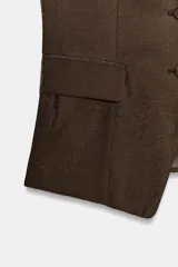 Chaleco marrón de corte sastre con escote recto, tirantes finos y cierre frontal con botones forrados a tono. Presenta bolsillos delanteros con solapa y un acabado con efecto brillo.