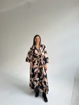Vestido largo negro con estampado de hojas en tonos beige y tostado, de corte amplio y fluido, con escote en V y botonadura frontal parcial. Cuenta con mangas largas y volados en el faldón.