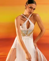 Vestido largo blanco tejido a mano en crochet, con escote halter cruzado y detalle de tejido en color dorado en el torso. Presenta aberturas laterales con flecos largos que caen desde la cintura.