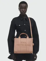 Cartera tipo tote de cuero granulado en color rosado, con diseño estructurado y correas dobles superiores. Incluye una correa ajustable y extraíble para llevar al hombro o cruzada. Presenta el logo 'THE TOTE BAG MARC JACOBS' grabado en el frente.