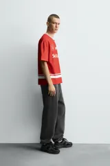 Camiseta de manga corta color rojo, corte boxy fit, con cuello pico ribeteado en blanco. Presenta un estampado frontal con el texto "Social Leagues", una estrella blanca y un escudo con el número "4". Tiene detalles de franjas blancas en el bajo y en los hombros.