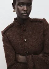 Chaqueta estructurada de tweed en color marrón, de corte recto con cuello redondo, manga larga y cierre frontal de botones. Presenta terminaciones deshilachadas y bolsillos frontales.