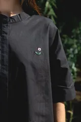 Camisa blanca de corte oversize y largo midi, confeccionada en algodón. Presenta cuello mao, cierre frontal oculto por tapeta y mangas cortas con dobladillo. Incluye un pequeño bordado de una flor en el pecho.