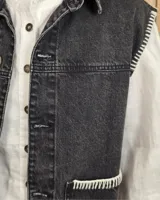 Chaleco de denim color marrón, confeccionado en 100% algodón, con bolsillos frontales amplios, cierre de botones y costuras decorativas bordadas en blanco en los bordes y los bolsillos.