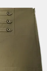 Shorts mini verde militar de tiro medio con aplicación de tiras horizontales y botones metálicos en el frente. Cuenta con cierre lateral de cremallera oculta.