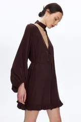 Vestido oversized corto de color marrón oscuro, con escote en V pronunciado y pañuelo haciendo juego que puede abrocharse en la nuca. Cuenta con botones forrados adelante, cinturón fino para atar, mangas globo largas de corte raglán con elástico en los puños y ribete de volados en el bajo.