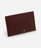 Billetera compacta de poliuretano color bordó, con diseño slim, ranuras para tarjetas en ambos lados y aplicación metálica frontal con las letras "STN".