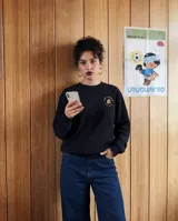 Buzo de cuello redondo color negro, con estampado circular en la espalda que presenta la palabra 'PRESENTE' en tipografía gótica de color beige y gris. Cuenta con puños y cintura acanalados.