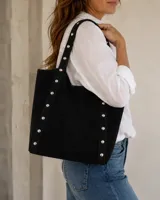Cartera tipo tote de símil gamuza color negro, con diseño amplio y detalle de tachas metálicas en las costuras y las asas de hombro.
