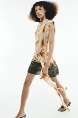 Vestido corto sin mangas de cuello redondo, con estampado tie-dye en tonos beige, naranja y verde oscuro. Presenta un lazo para atar en la cintura y cierre trasero con cremallera oculta.