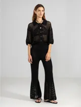 Camisa corta de punto abierto con cuello tipo polo, cierre frontal de botones y mangas tres cuartos. Presenta un diseño calado y un botón decorativo en forma de flor.