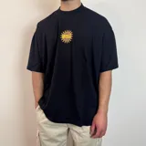 Remera negra de corte oversize con cuello redondo alto y estampado gráfico de un sol con el texto MIDWY STUDIO en el centro del pecho.