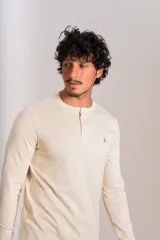 Remera de manga larga tipo Henley, color beige, con cuello redondo y tapeta con botones. Presenta un pequeño logo bordado en el pecho.