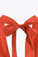 Top de color naranja vibrante con cuello halter que se anuda en la espalda, creando un escote drapeado o fluido en el frente. El bajo del top tiene un corte en pico con tiras finas colgantes.