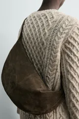 Bolso de hombro de piel con acabado serraje, color gris oscuro, con forma curva o de media luna. Presenta costuras visibles en el centro y un asa fina para llevar al hombro.