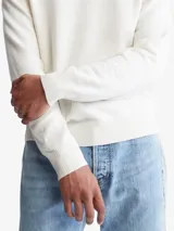 Sweater de punto liso color blanco hueso, con cuello redondo y puños y dobladillo en punto canalé. Presenta un pequeño logo bordado en el pecho.