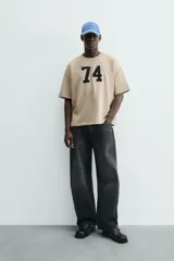 Sudadera de manga corta color beige, corte relajado (relaxed fit) con cuello redondo y número "74" bordado en negro en el frente.