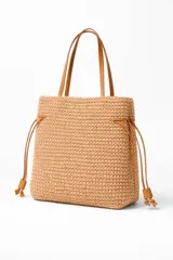 Bolso tipo tote bag color beige, tejido a crochet, con asas de cuero marrón y cordones laterales ajustables.