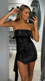 Vestido corto strapless de tela satinada negra, con diseño ajustado y fruncido lateral. Presenta un detalle de caída asimétrica en uno de los costados.