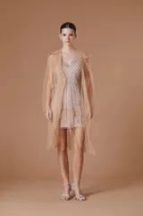 Trench ligero de tul color nude, con diseño abierto, solapas clásicas y mangas largas. Presenta una caída fluida y transparente, ideal para superposiciones elegantes.