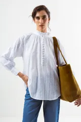 Blusa de algodón blanco con diseño de rayas verticales y textura de pequeños puntos en relieve. Presenta cuello alto con volados, cierre frontal con botones forrados y mangas largas abullonadas con puños ajustados.