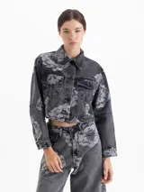 Chaqueta cropped de denim con lavado gris y estampado floral en contraste. Presenta cuello clásico, mangas largas, bolsillos frontales con tapa y cierre mediante broches metálicos.