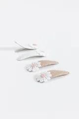 Pack de cuatro broches para el pelo tipo clip, con diseño de flor de tela aplicada en la parte superior. Dos broches son de color blanco liso y dos son de color beige con estampado de pequeñas estrellas blancas.