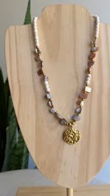 Collar corto combinado con cuentas de nácar en tonos marrones y beige, cuentas de piedra reconstruida blanca y apliques de acero quirúrgico dorado. Incluye un dije redondo dorado con diseño de estrella.