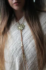 Corbatín con dije de bronce con forma de flor y tiras de cuero sintético marrón con detalles metálicos dorados.