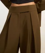 Pantalón de sastrería corte barrel, color marrón oscuro, con pliegues delanteros, cintura con cierre desplazado y piernas anchas.
