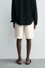 Bermuda de corte relajado color beige claro, confeccionada en tejido de algodón y lino. Presenta cintura con cordón ajustable del mismo tejido, bolsillos delanteros y bolsillos traseros de vivo. Cierre frontal con cremallera y botón.