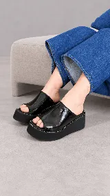 Zueco de plataforma corrida modelo Dakota, confeccionado en material sintético efecto charol de color negro, con diseño abierto en la punta y costuras blancas contrastantes en el borde de la suela.