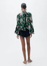Blusa de gasa con estampado floral en tonos rosa, verde y negro. Tiene cuello alto, mangas largas con volantes y cierre de lazo en la parte posterior.