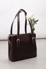 Cartera de hombro estructurada color negro, confeccionada en 100% cuero vacuno con textura en relieve tipo reptil. Cuenta con asas largas ajustables mediante hebillas metálicas plateadas, cierre superior y el logo de la marca grabado en la parte frontal.