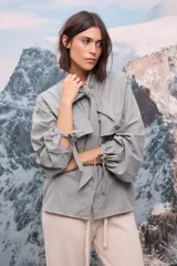 Camisa de tela afelpada color verde grisáceo, con cuello con lazo anudado, mangas largas abullonadas con detalles de lazos en los puños y cierre frontal con botones.