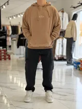 Canguro oversize beige con capucha y cordones ajustables, bolsillo tipo canguro y pequeño bordado en el pecho.