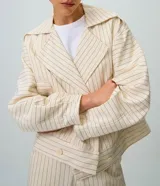 Tapado corto tipo trench coat confeccionado en sarga de algodón, de color beige con estampa de rayas finas verticales y horizontales. Presenta cuello solapa, mangas largas, bolsillos laterales con ribete y cierre frontal cruzado con doble botonadura.