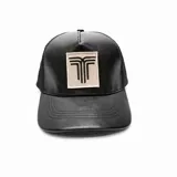 Gorra tipo trucker de color negro, confeccionada en cuero sintético en la parte frontal y visera, con paneles traseros de malla. Presenta un parche rectangular en el centro con un diseño gráfico bordado en color negro sobre fondo beige.