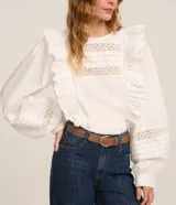 Blusa blanca de tricolina con diseño romántico, cuello redondo, mangas largas abullonadas y detalles de volados verticales en el frente. Presenta aplicaciones de encaje tipo entremedios en el pecho y en las mangas.