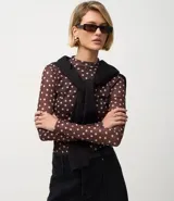 Blusa de manga larga ajustada al cuerpo, confeccionada en tul marrón con estampado de lunares blancos. Presenta cuello redondo y un cárdigan negro cruzado sobre los hombros.