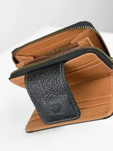 Billetera compacta de cuero negro con diseño combinado de textura granulada y efecto crocco brillante. Cuenta con cierre perimetral, solapa frontal con broche y logo grabado, e incluye una correa de muñeca extraíble.